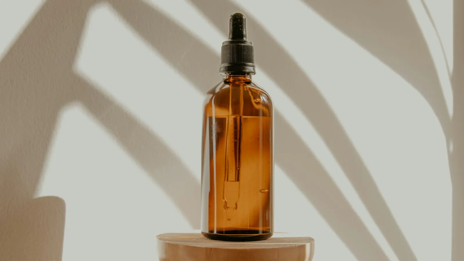 An amber dropper bottle on a wooden pedestal lit by morning light through palm shadows