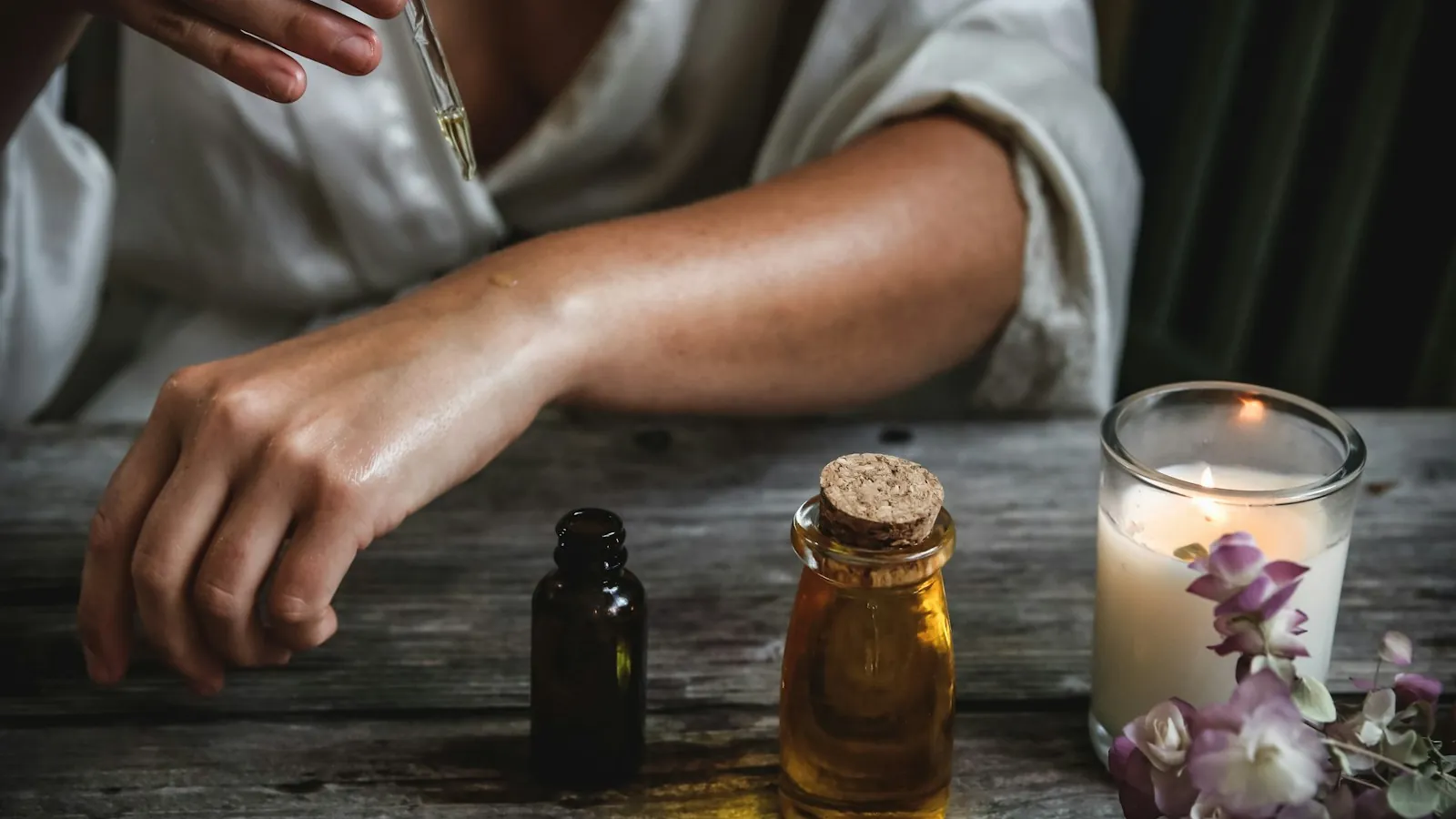 Hand applying essential oil from a dropper next to amber and clear glass bottles, a candle, and dried flowers on a wood surface