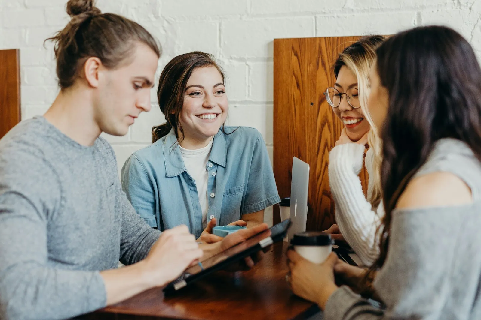 Four friends sitting around a cafe table, three looking together at a tablet